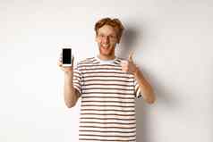 技术电子商务概念年轻的男人。红色的头发显示竖起大拇指空白智能手机屏幕推荐应用程序白色背景