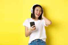 很酷的亚洲女孩唱歌听音乐无线耳机持有智能手机手站黄色的背景