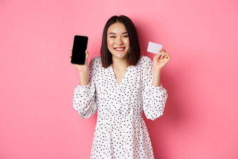 快乐的朝鲜文女人显示智能手机屏幕信贷卡支付互联网订单展示在线购物应用程序站粉红色的背景