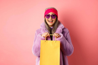 时尚亚洲高级女人购物太阳镜时尚的冬天衣服持有纸袋商店微笑快乐相机粉红色的背景
