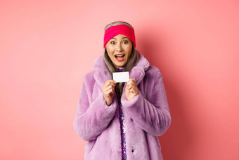 购物时尚概念美丽的亚洲中年女人显示塑料信贷卡微笑惊讶时尚的衣服粉红色的背景