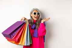 时尚的高级亚洲女人太阳镜购物假期出售持有纸袋微笑站白色背景