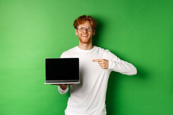 快乐红色头发的人的家伙眼镜白色长袖t恤指出手指空白移动PC屏幕微笑站绿色背景