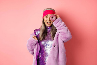 食物时尚概念美丽的亚洲高级女人迪斯科衣服人造皮毛外套甜甜圈洞惊讶脸站快乐粉红色的背景