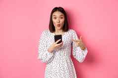 在线购物美概念惊讶亚洲女人指出手指移动电话推荐智能手机应用程序站粉红色的背景