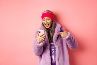 在线购物时尚概念很酷的亚洲<strong>高级</strong>女人检查促销活动移动电话指出手指智能手机屏幕微笑很高兴粉红色的背景