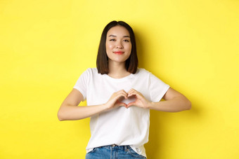 美丽的亚洲女人显示爱心手势微笑相机站黄色的背景概念情人节一天浪漫