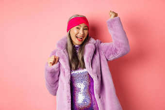 购物概念快乐的高级亚洲女人跳舞有趣的穿时尚的<strong>紫色</strong>的人造皮毛<strong>外套</strong>衣服赢得奖微笑站粉红色的背景