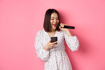 可爱的亚洲女人阅读<strong>歌词</strong>智能手机唱歌麦克风站时尚的衣服粉红色的背景