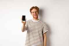 技术电子商务概念快乐年轻的红色头发的人男人。眼镜显示空白智能手机屏幕微笑满意站白色背景