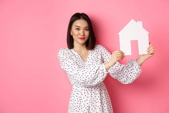 真正的房地产美丽的亚洲女人展示纸房子模型相机自信广告首页出售站粉红色的背景