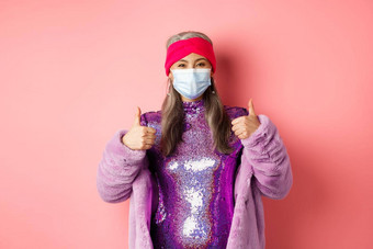 冠状病毒购物概念时尚的亚洲祖母呼吸器闪闪发光的紫色的衣<strong>服人</strong>造皮毛外套显示竖起大拇指推荐商店脸面具