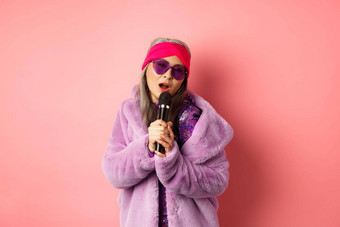 时尚概念时尚的高级亚洲女人唱歌<strong>卡拉ok</strong>执行阶段麦克风穿时尚的太阳镜紫色的人造皮毛外套粉红色的背景