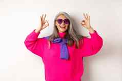 时尚的现代亚洲祖母太阳镜粉红色的毛衣微笑快乐显示迹象批准很酷的白色背景
