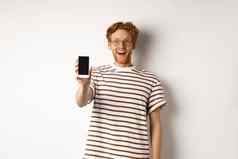 技术电子商务概念快乐年轻的红色头发的人男人。眼镜显示空白智能手机屏幕相机惊讶站白色背景