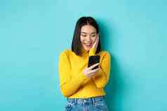 图像快乐亚洲女人阅读消息移动电话屏幕微笑闲谈，聊天智能手机应用程序站蓝色的背景