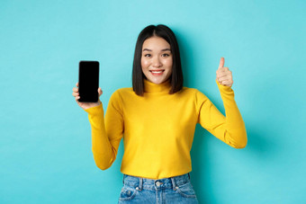 技术人概念快乐的亚洲女孩黄色的毛衣显示空白智能手机屏幕拇指演示在线提供站蓝色的背景
