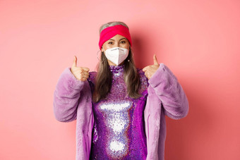 科维德流感大流行时尚概念快乐的亚洲<strong>高级</strong>女人聚会冠状病毒呼吸器显示竖起大拇指推荐穿脸面具粉红色的背景