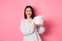 浪漫的亚洲女人可爱的衣服持有盒子礼物微笑快乐相机站现在粉红色的背景