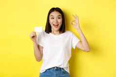 年轻的亚洲女人休闲白色t恤显示塑料信贷卡手势推荐银行微笑相机黄色的背景