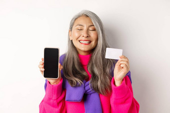 在线购物快乐很高兴亚洲<strong>高级</strong>女人微笑快乐显示空白智能手机屏幕塑料卡购买互联网白色背景
