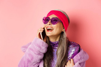时尚美丽的亚洲高级女人时尚的太阳镜头巾笑会说话的移动电话粉红色的背景