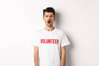 震惊了困惑男人。志愿者t恤盯着相机皱着眉头不高兴站白色背景