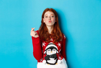 冬天假期圣诞节夏娃概念可爱的红色头发的人女孩圣诞节毛衣显示心标志皱<strong>纹嘴唇</strong>吻站蓝色的背景
