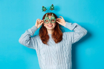 冬天假期圣诞节销售概念美丽的红色头发的人女模型庆祝一年穿有趣的聚会，派对头巾眼镜微笑愚蠢的蓝色的背景