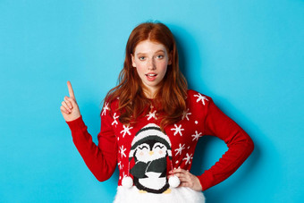 冬天假期圣诞节夏娃概念时髦的红色头发的人女孩圣诞节毛衣指出上左角落里盯着相机提示促销提供蓝色的背景