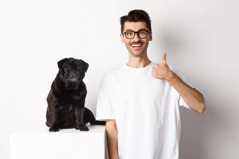 图像狗老板可爱的黑色的哈巴狗相机男人。显示翘拇指批准推荐站白色背景