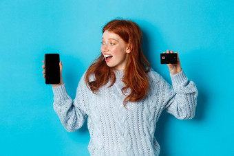 兴奋红色头发的人女孩显示移动电话屏幕信贷卡展示在线<strong>商店应用</strong>程序站蓝色的背景