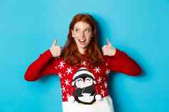 冬天假期圣诞节夏娃概念美丽的红色头发的人女孩圣诞节毛衣庆祝一年显示拇指上左角落里蓝色的背景