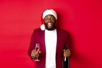 圣诞节聚会，派对假期概念兴奋非洲美国男人。圣诞老人他庆祝一年持有瓶玻璃香槟站红色的背景