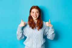 快乐的青少年女孩红色的头发显示拇指批准赞美手势站蓝色的背景