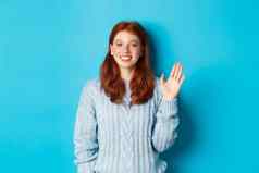 友好的红色头发的人十几岁的女孩挥舞着手手势微笑站蓝色的背景