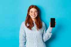 微笑女模型红色的头发显示智能手机屏幕持有电话展示应用程序站蓝色的背景