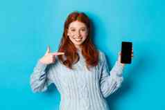 满意红色头发的人女孩指出电话屏幕显示智能手机应用程序在线促销微笑站毛衣蓝色的背景