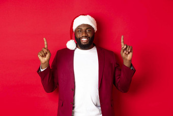 圣诞节聚会，派对假期概念英俊的非洲美国男人。圣诞老人他微笑指出手指显示广告站红色的背景