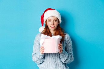 冬天假期圣诞节夏娃概念可爱的红色头发的人女孩穿圣诞老人他持有一年礼物相机站蓝色的背景