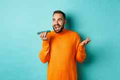 快乐男人。会说话的扬声器手势记录的声音消息移动电话站橙色毛衣光蓝色的背景