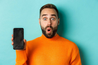 特写镜头英俊的有胡子的的家伙橙色毛衣显示智能手机屏幕微笑显示促销在线绿松石背景