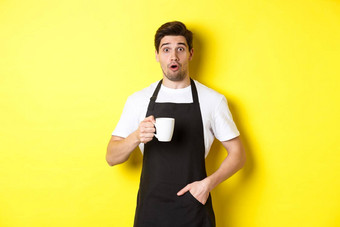 咖啡师持有咖啡杯子惊讶站黑色的围裙咖啡馆统一的黄色的背景