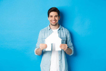 保险抵押贷款真正的房地产概念微笑年轻的男人。持有房子模型搜索公寓租金站蓝色的背景