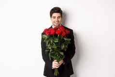 图像英俊的的男朋友黑色的西装持有花束红色的玫瑰微笑日期站白色背景