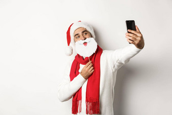 圣诞节聚会，派对庆祝活动概念年轻的男人。采取自拍有趣的白色胡子圣诞老人面具他摆姿势照片移动电话工作室背景
