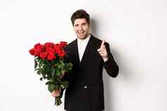 图像英俊的浪漫的的家伙黑色的西装持有花束玫瑰指出相机祝贺假期站白色背景