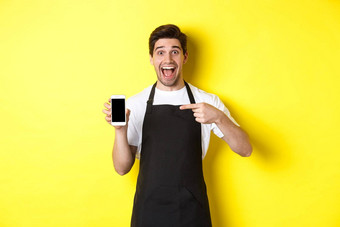 英俊的咖啡师黑色的围裙指出手指移动屏幕显示应用程序微笑站黄色的背景