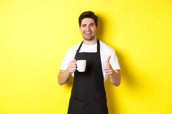 英俊的咖啡师黑色的围裙持有咖啡杯指出手指邀请访问咖啡馆站黄色的背景