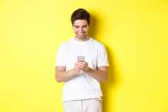 年轻的男人。阅读文本消息智能手机移动电话屏幕微笑站白色t恤黄色的背景